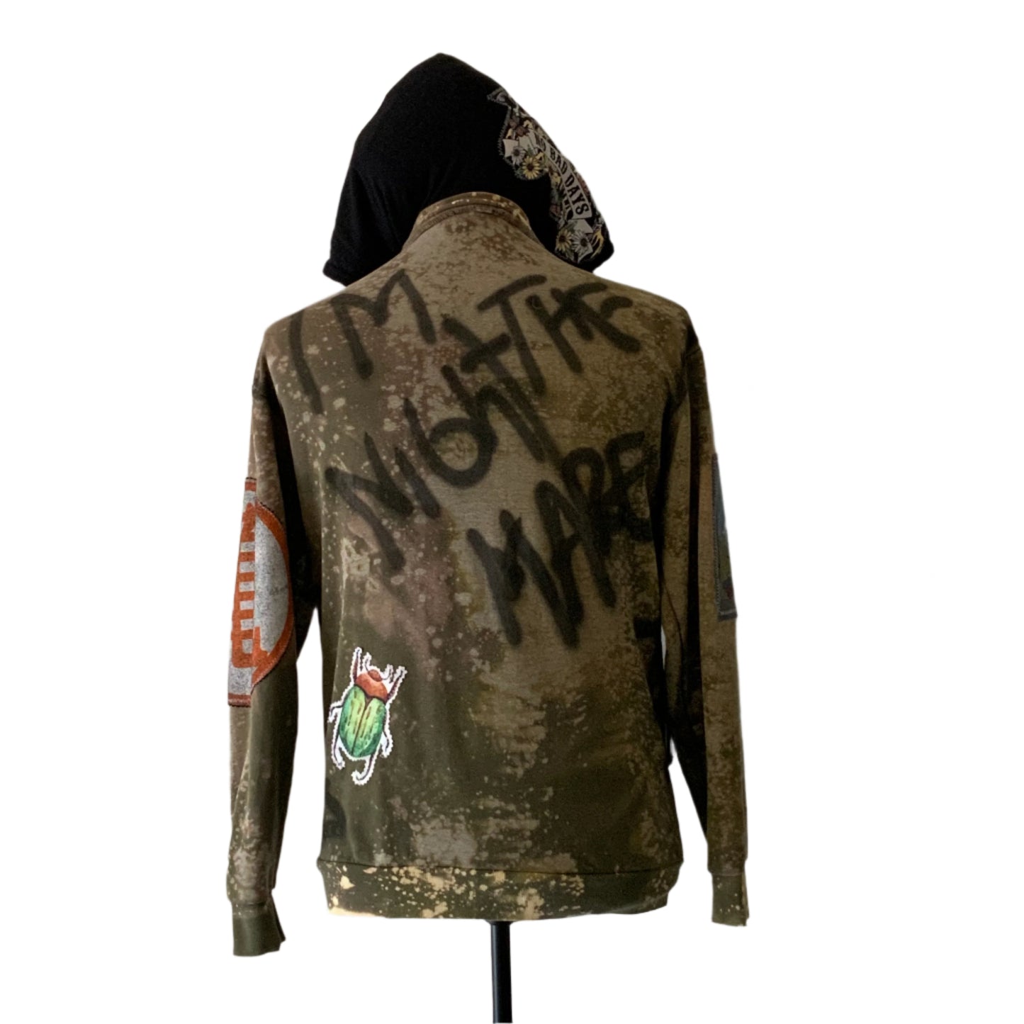 Custom army-green hoodie with bleach distressing, spray-painted I'M NOT THE MARE, colorful patches, and graffiti design. Represents bold, unique style from DEVIOUS SAINT.