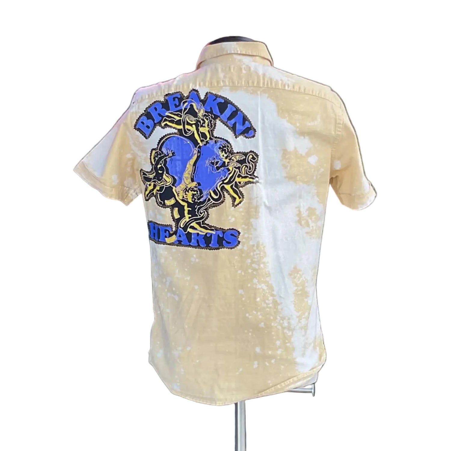Yellow cotton shirt featuring cherub graphic and BREAKIN’ HEARTS text, capturing DEVIOUS SAINT's unique, artistic style. Displayed on mannequin with foliage backdrop.