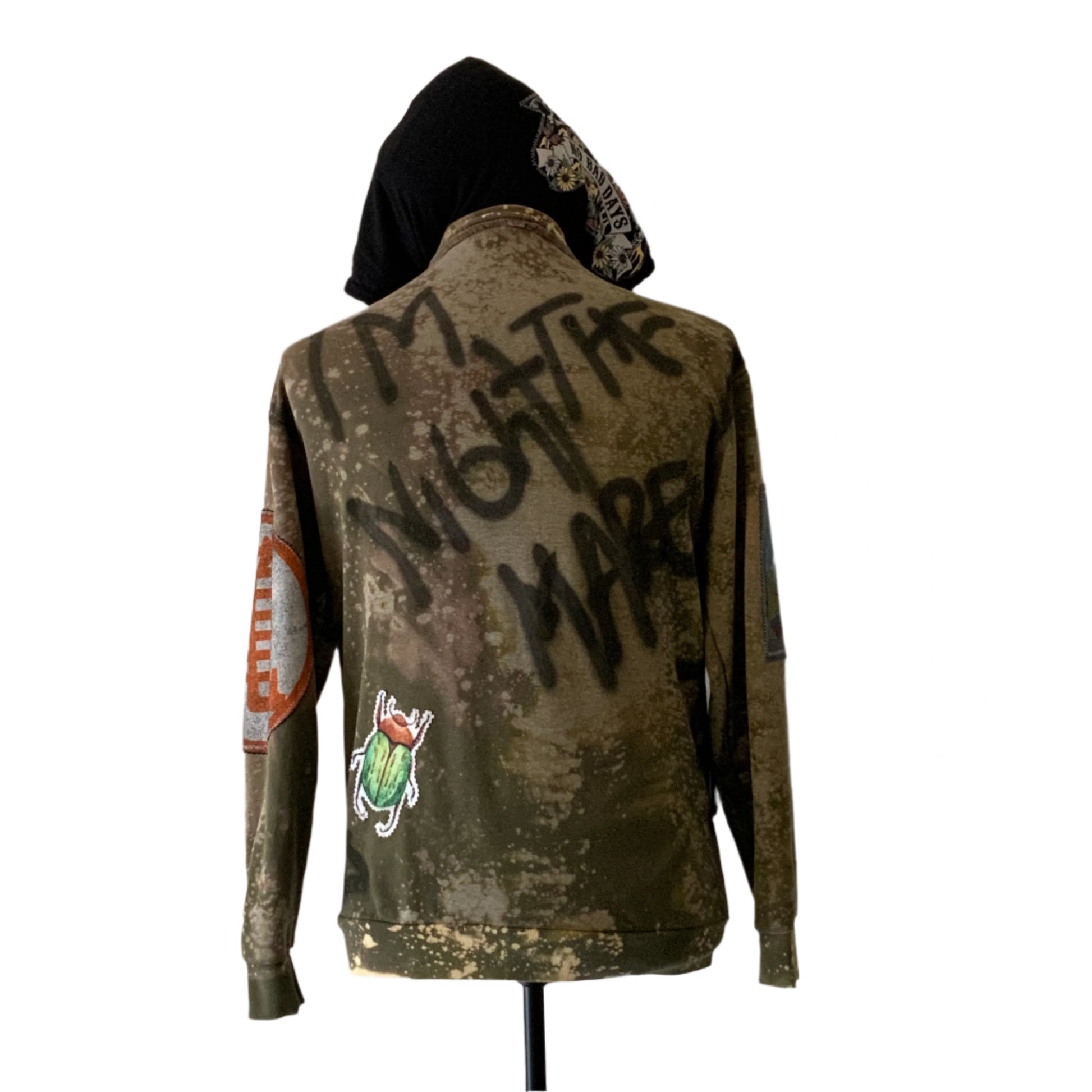 Custom army-green hoodie with bleach distressing, spray-painted I'M NOT THE MARE, colorful patches, and graffiti design. Represents bold, unique style from DEVIOUS SAINT.