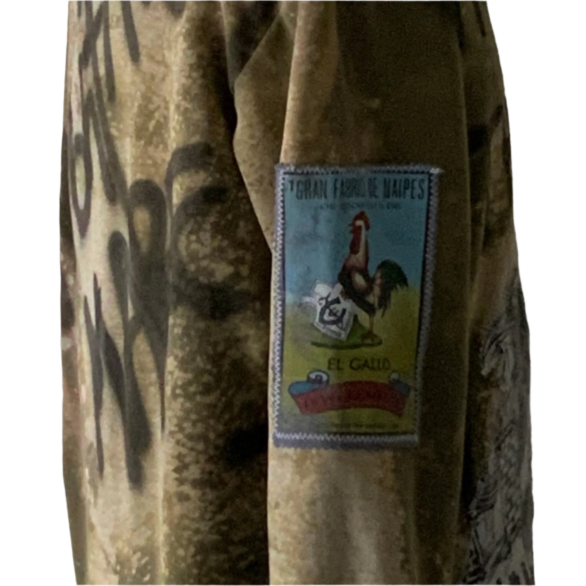 Camouflage-style hoodie sleeve with lotería-style patch featuring a rooster, highlighting the bold, unique style of the I’M NOT THE DREAM collection by DEVIOUS SAINT.