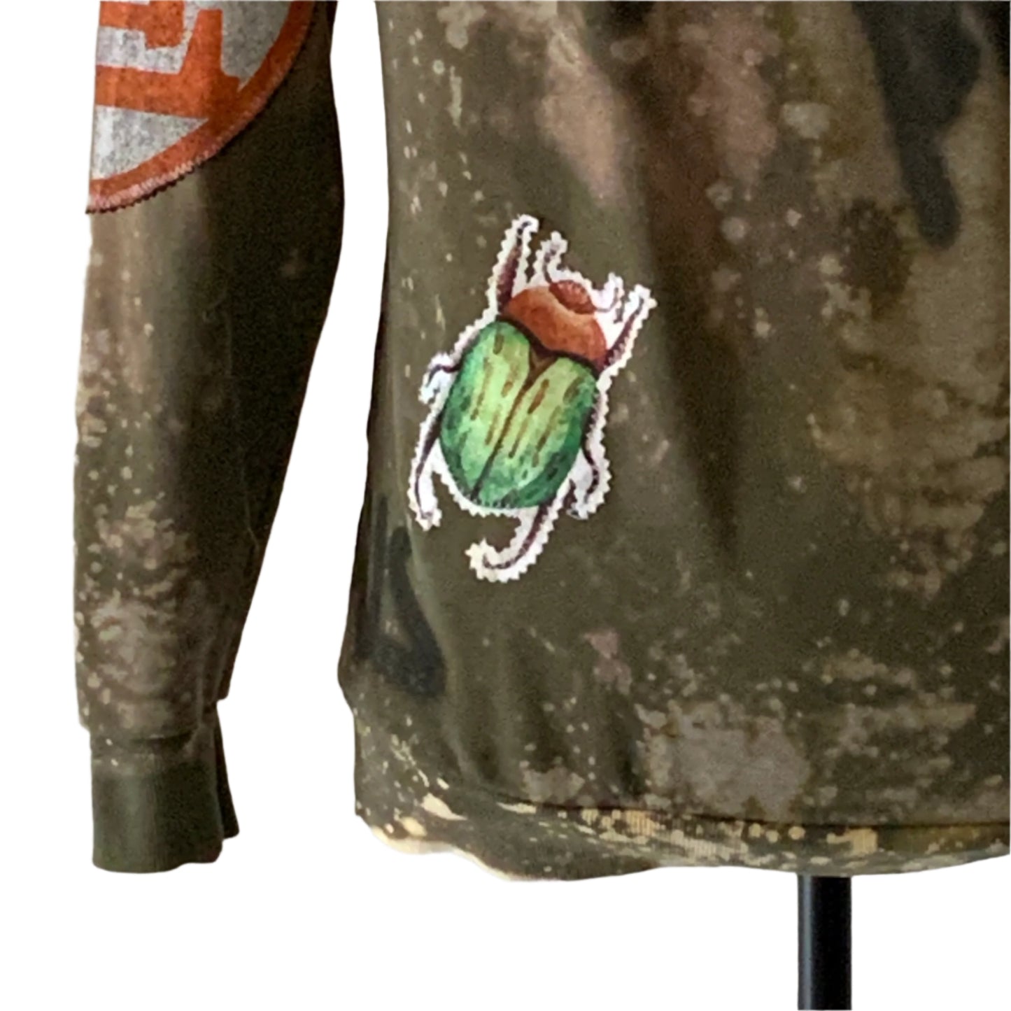 Olive-green long-sleeve shirt featuring bleach-splatter design and embroidered patches, including a green scarab beetle and orange graphic on sleeve.