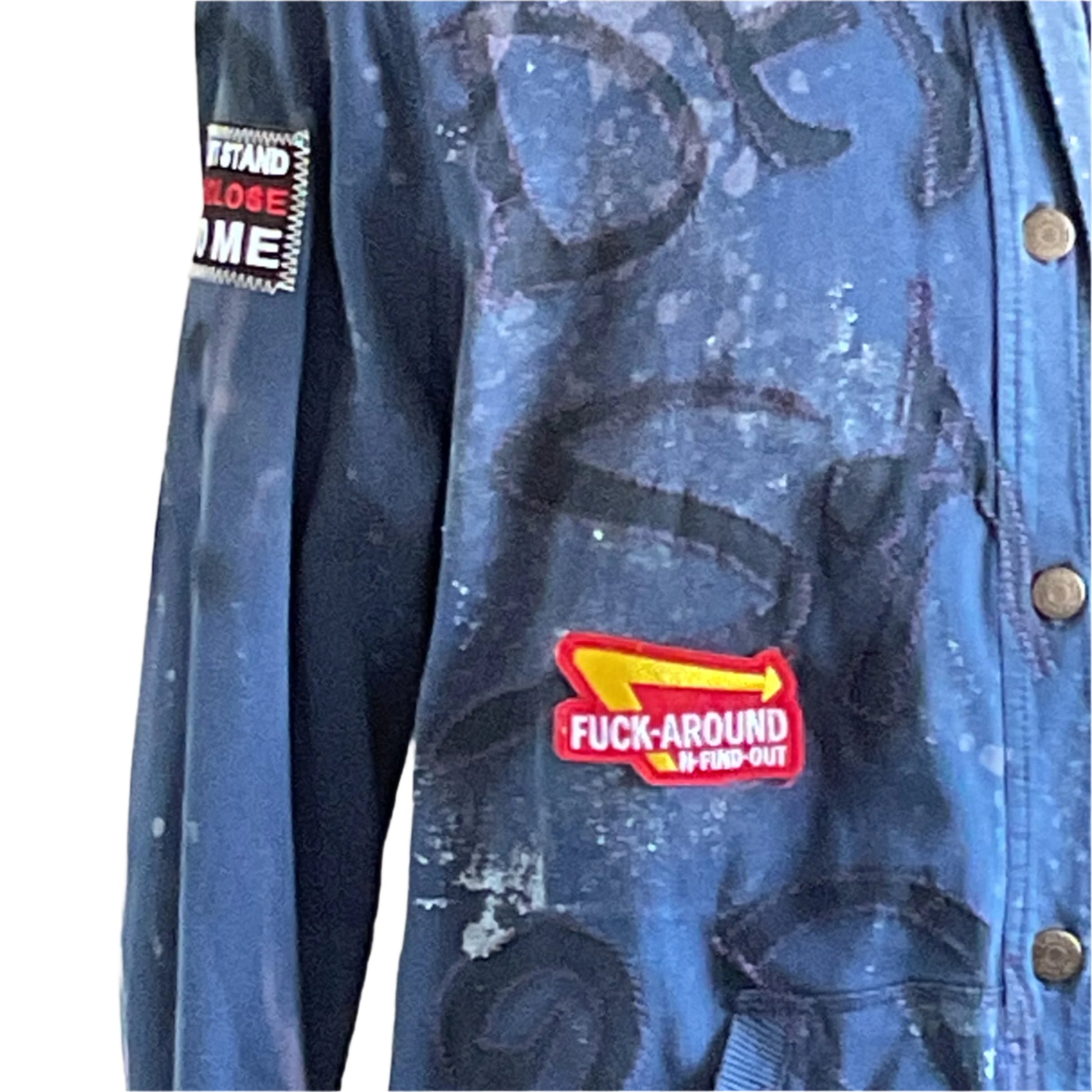 Close-up of TROUBLE MAKER jacket featuring yellow-arrow patch with FUCK-AROUND & FIND-OUT, brass snap buttons, and black swirl markings, embodying Devious Saint's rebellious style.