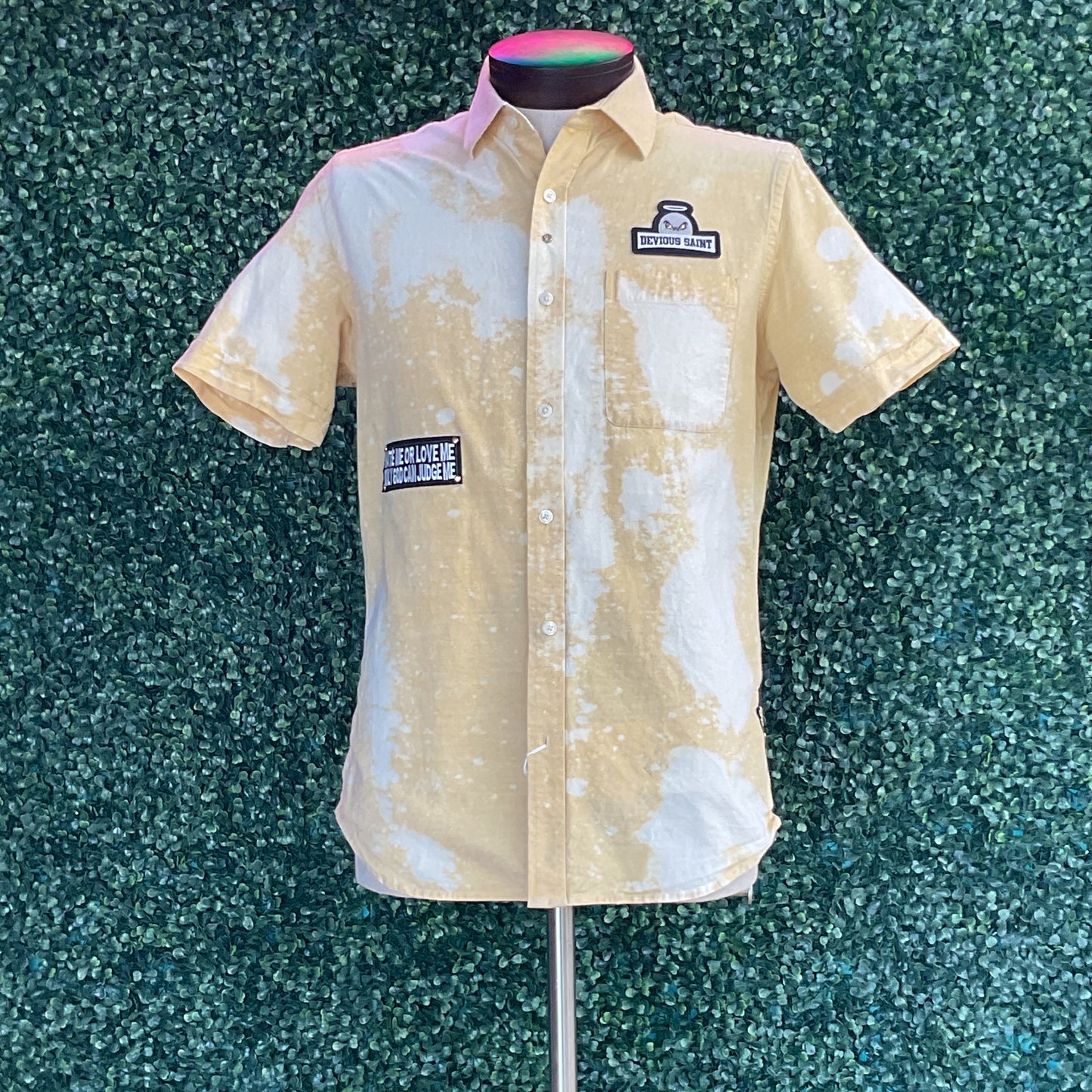 Men’s short-sleeve button-down shirt on a mannequin, featuring a washed-out pattern, chest pocket with DEVIOUS SAINT patch, and bold design.