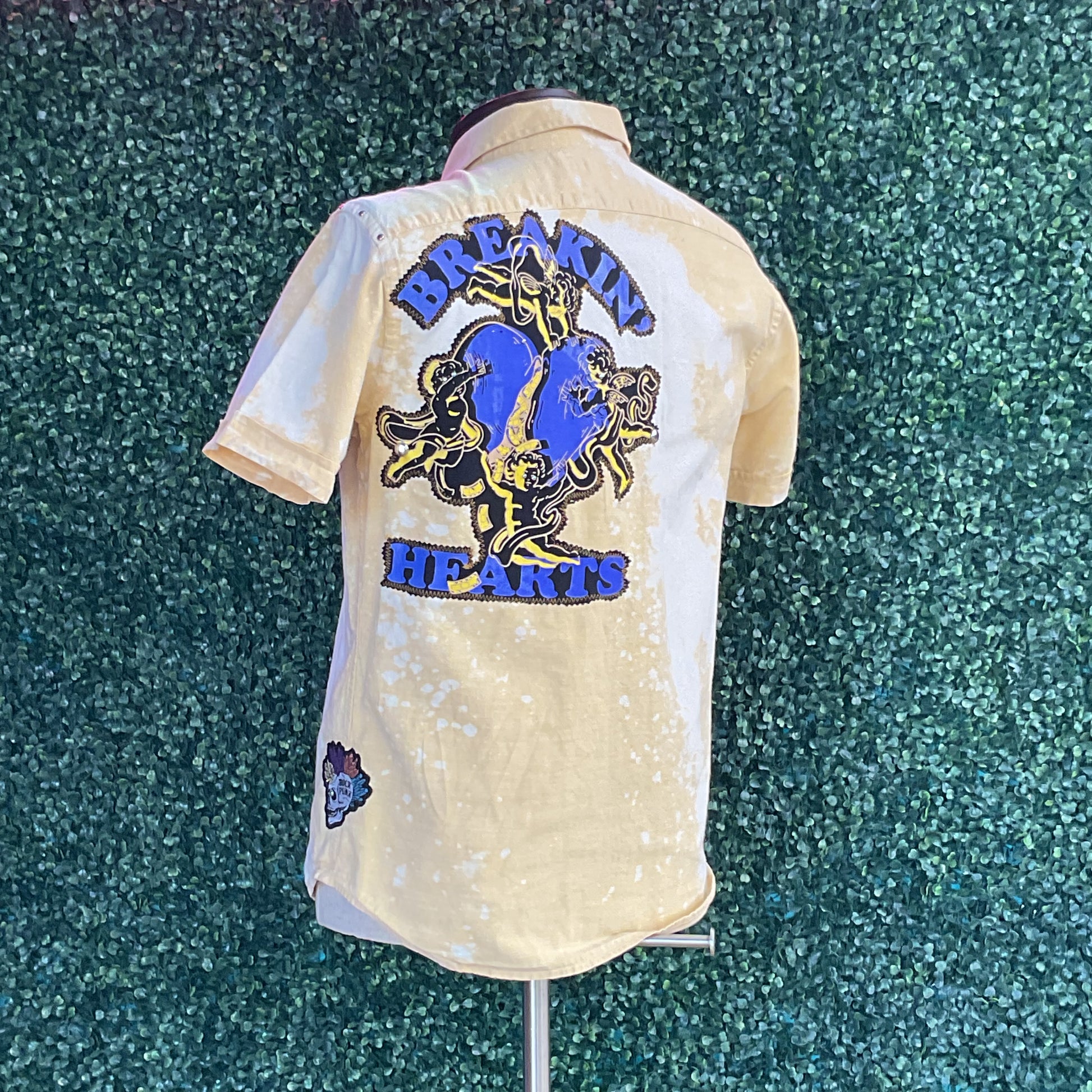 Yellow cotton button-up shirt featuring a large blue heart graphic with cherubs and BREAKIN’ HEARTS text, displayed on a mannequin, capturing bold, distinctive style.